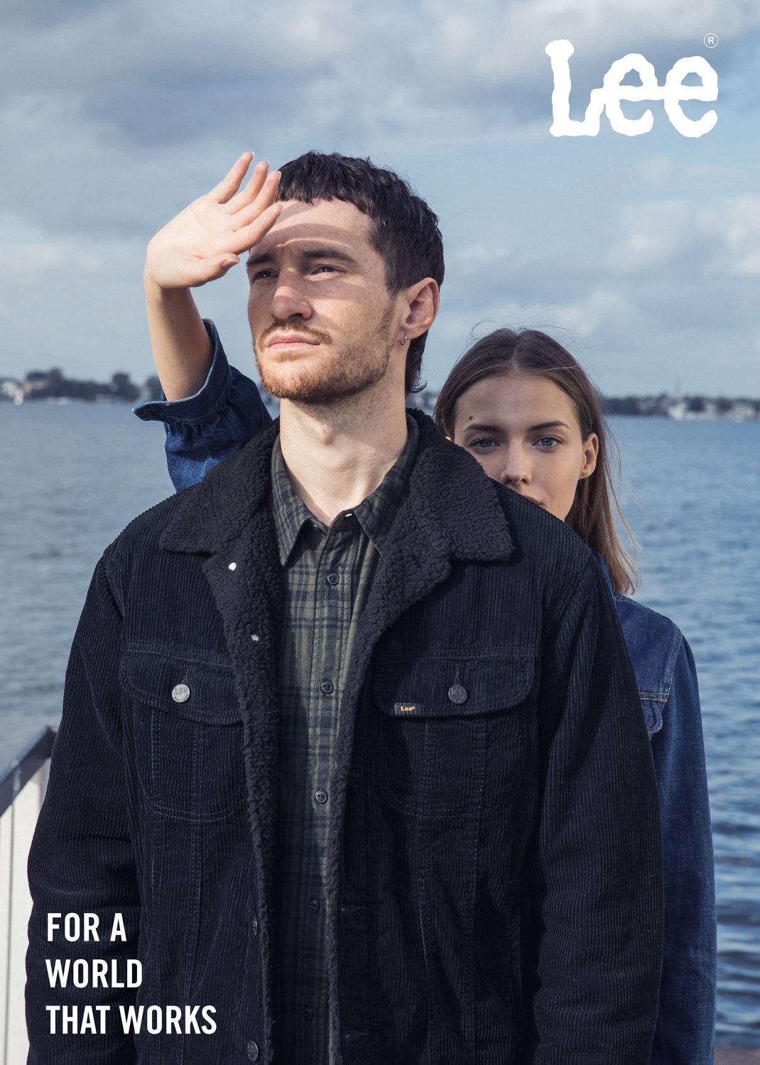 Two young adults, a man and a woman, outdoors near a body of water on a cloudy day. The man is in front, wearing a black jacket, looking into the distance while shielding his eyes from the sun. The woman stands behind him, partially visible, looking 