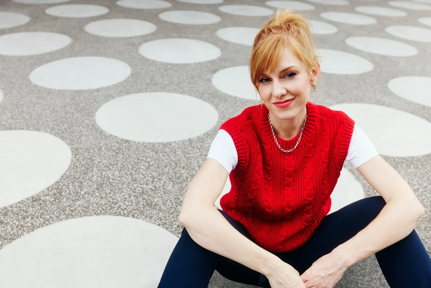 A young woman with red hair in a ponytail, wearing a red vest over a white t-shirt, sitting on a textured surface with large white circles, smiling and looking at the camera.