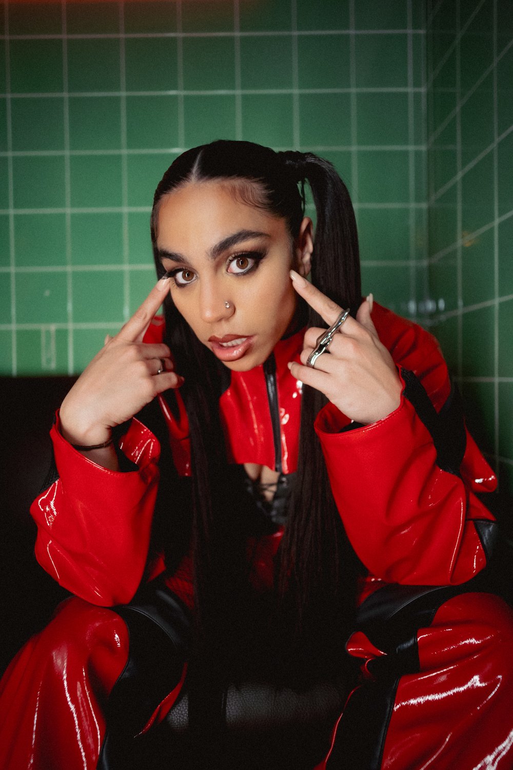 Young woman with dark hair in pigtails, wearing red and black vinyl outfit, sitting against green tiled wall, pointing to her temples with both index fingers.
