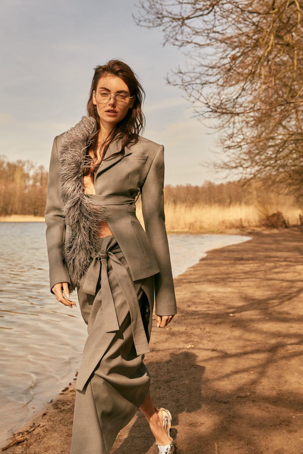 A woman wearing a stylish gray suit with fur detail walks along a riverbank during sunset.