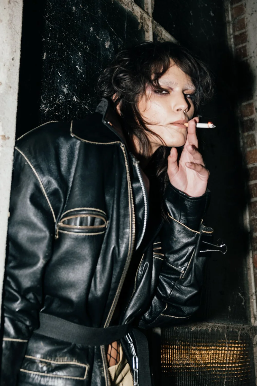 A woman with dark, messy hair and heavy makeup, wearing a black leather jacket, smokes a cigarette in a gritty, industrial setting with dark brick and black tiled walls.