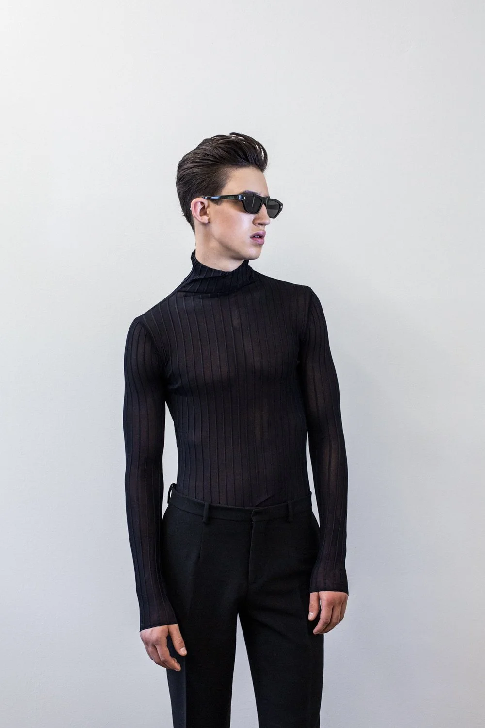 A person with dark hair styled in a pompadour, wearing black sunglasses, a black ribbed turtleneck, and black pants, standing against a plain light-colored wall.