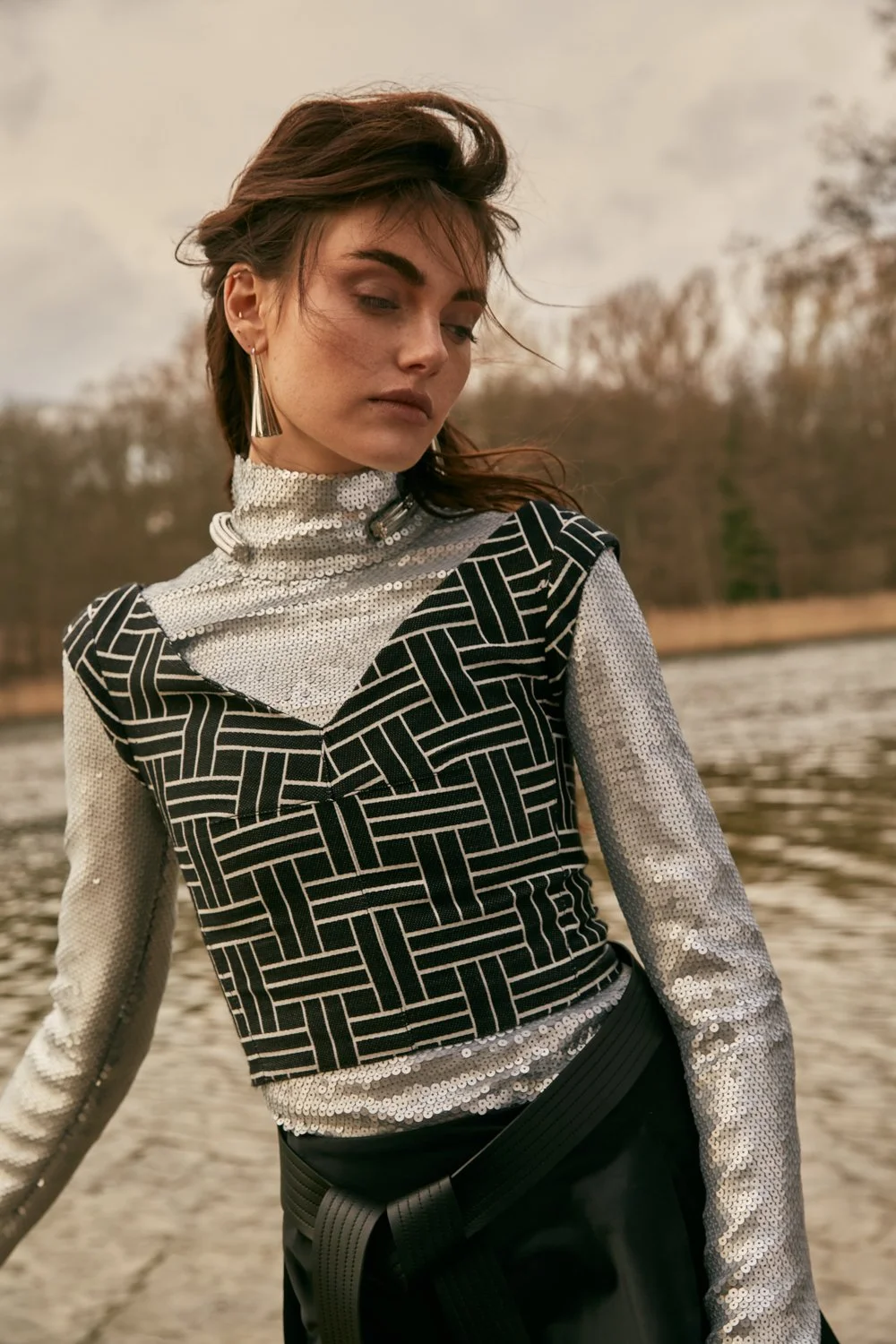 Woman with brown hair wearing a patterned black and white top with sequin sleeves, standing outdoors near a body of water with trees in the background.