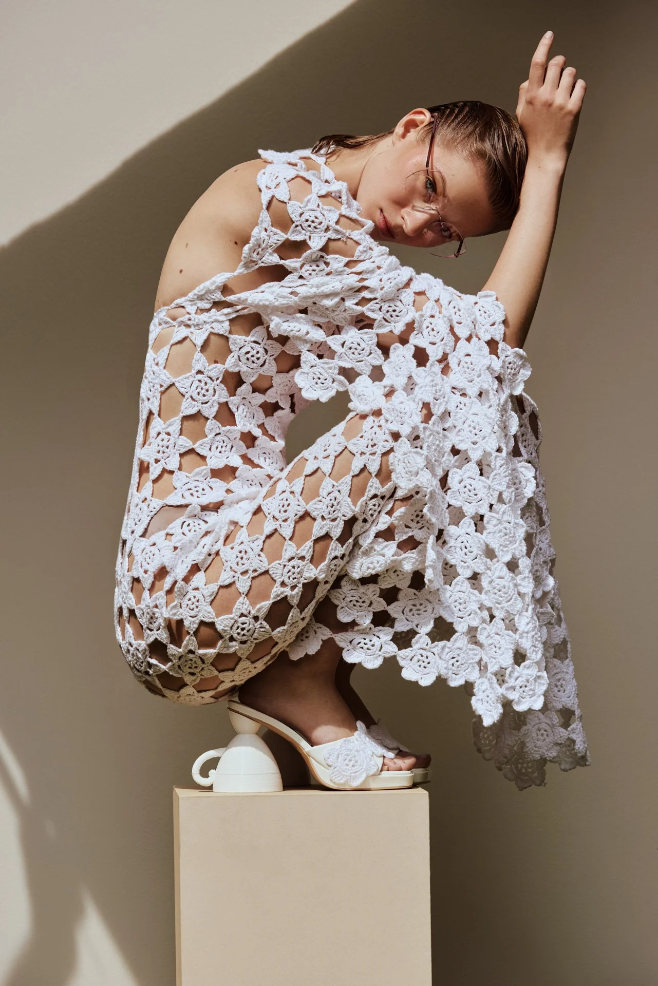 A woman with glasses and wet hair, dressed in a white crochet outfit, is squatting on a pedestal against a plain background, looking at the camera.