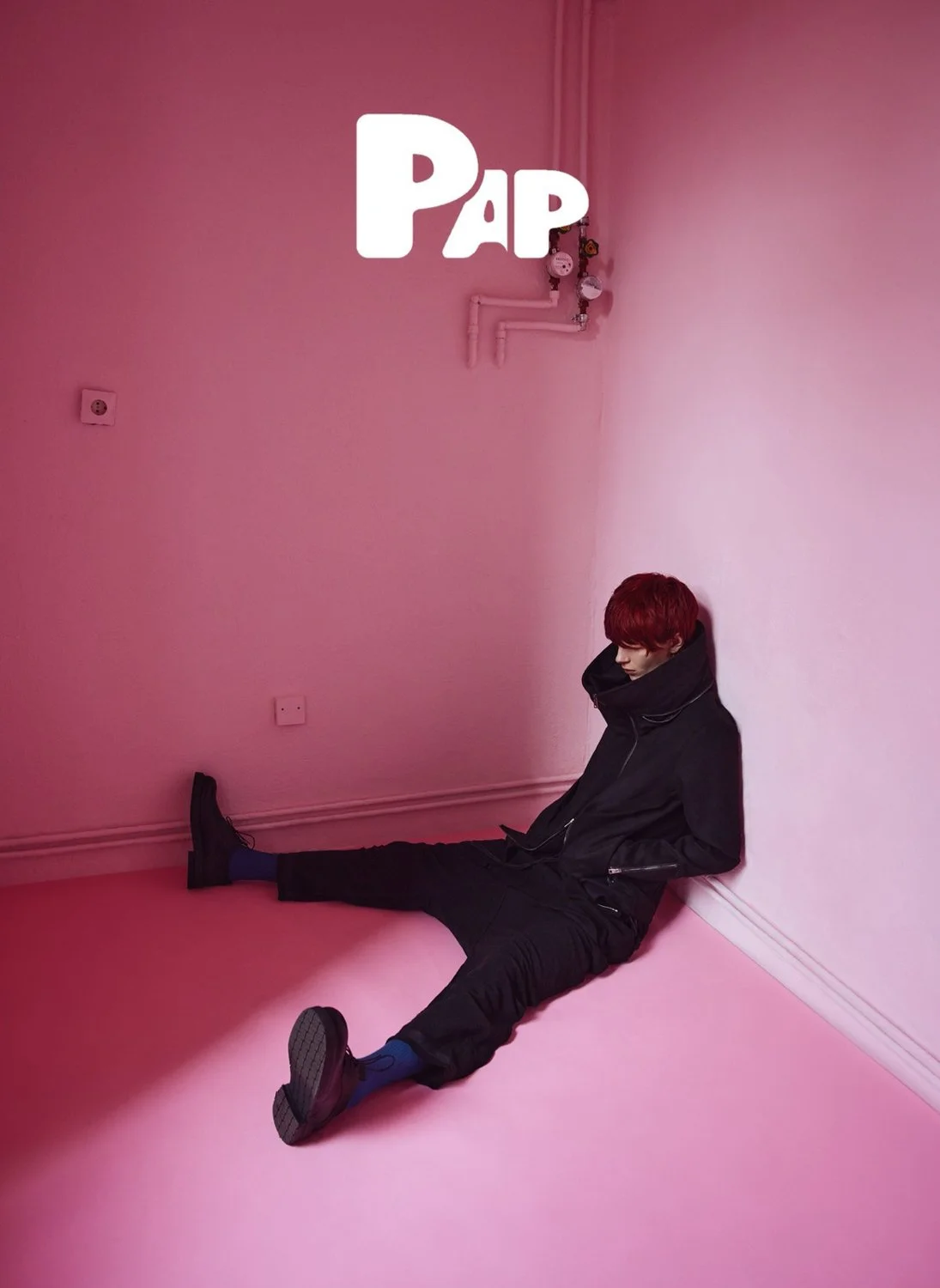 A person with red hair, dressed in black, sitting on the pink floor with their back against the pink wall, with the word 'PAP' written in white over the image.