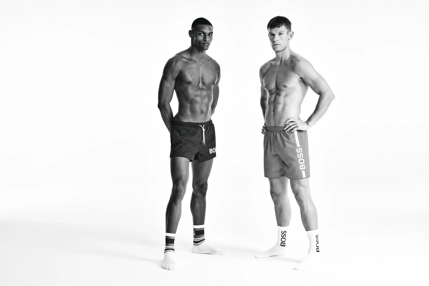 Two shirtless men modeling BOSS brand shorts and socks in a studio with a plain white background.
