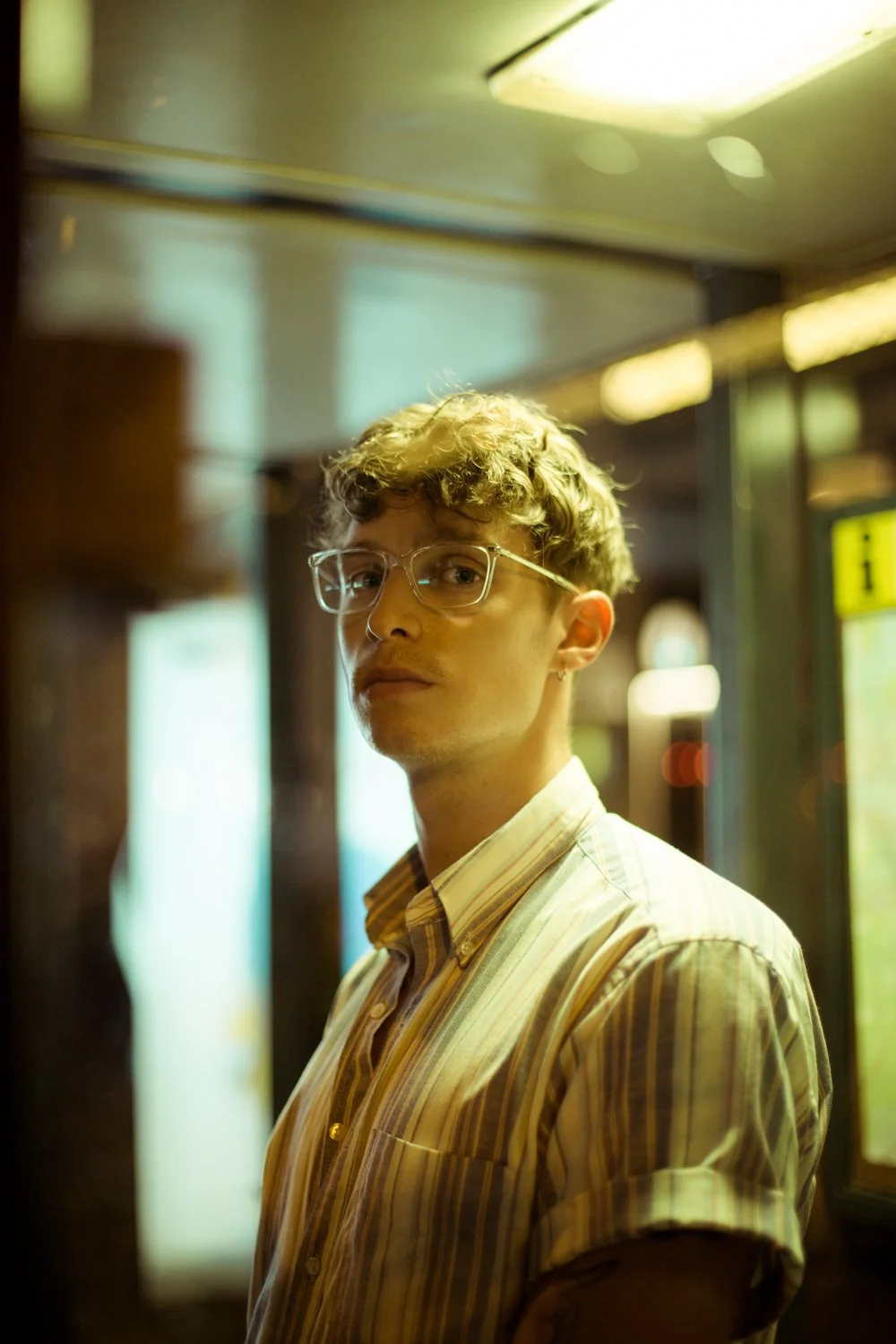 Young man with curly hair and glasses looking at camera in a dimly lit indoor space with a bright sign in the background.