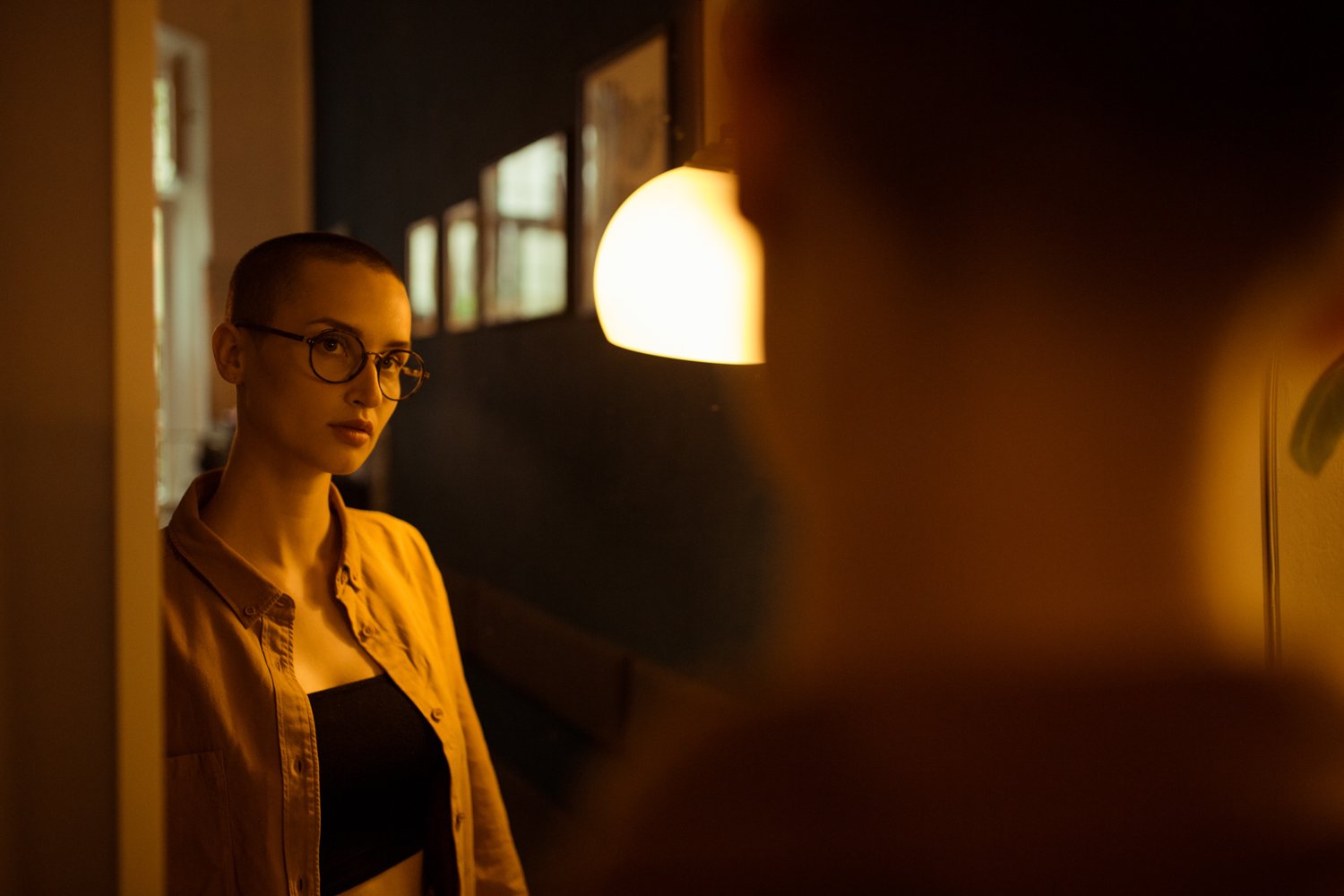Woman with short hair and glasses standing indoors near a mirror, illuminated by warm yellow light, with framed pictures on the dark wall behind her.