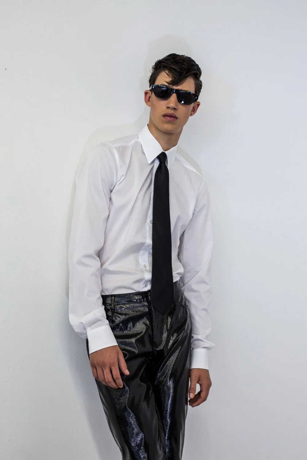 Young man in white dress shirt, black tie, black sunglasses, and shiny black pants leaning against a white wall.