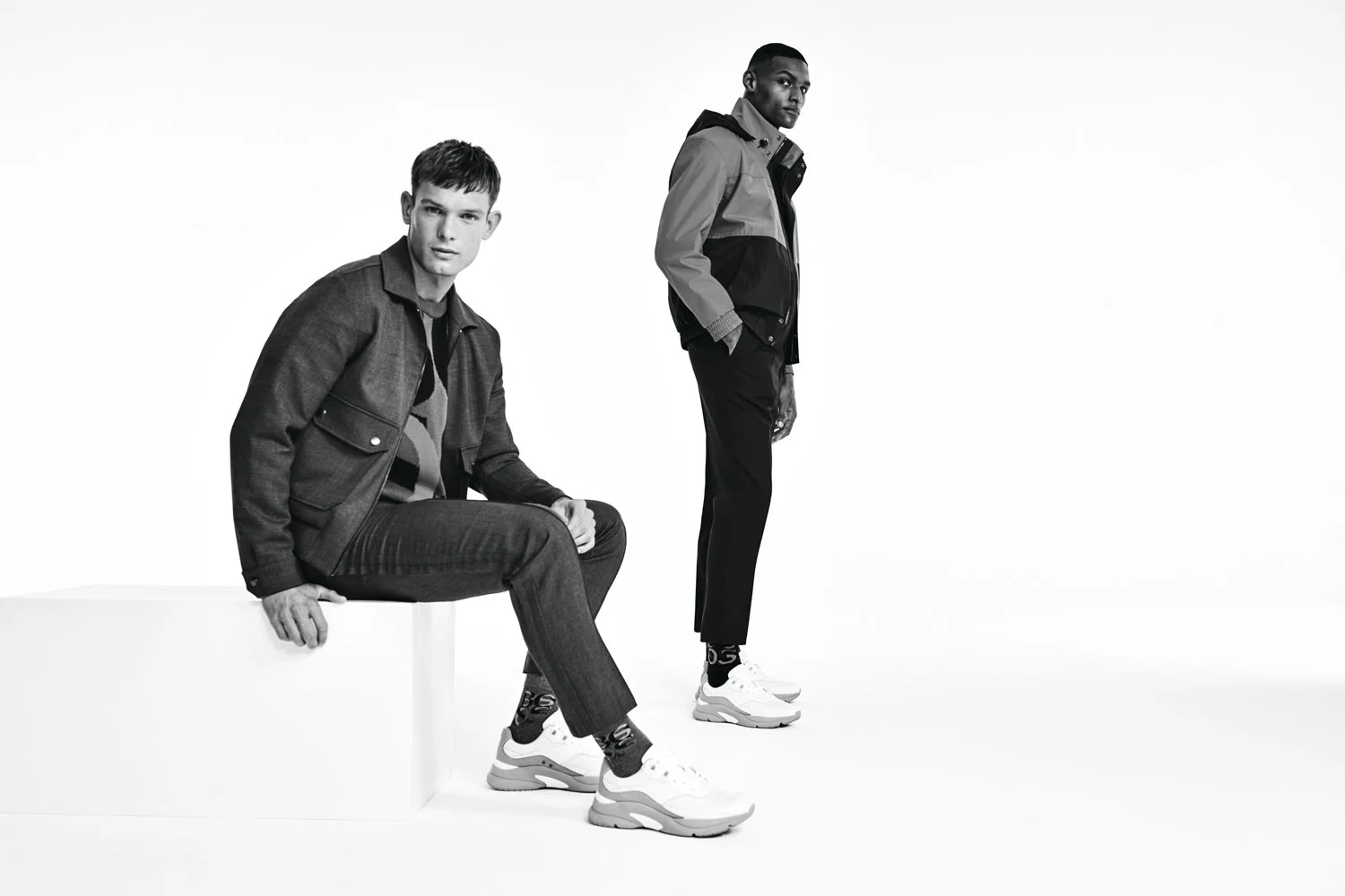 Two men with athletic builds wearing casual sportswear in black and white studio shot. One man is sitting on a white block, the other standing beside him.