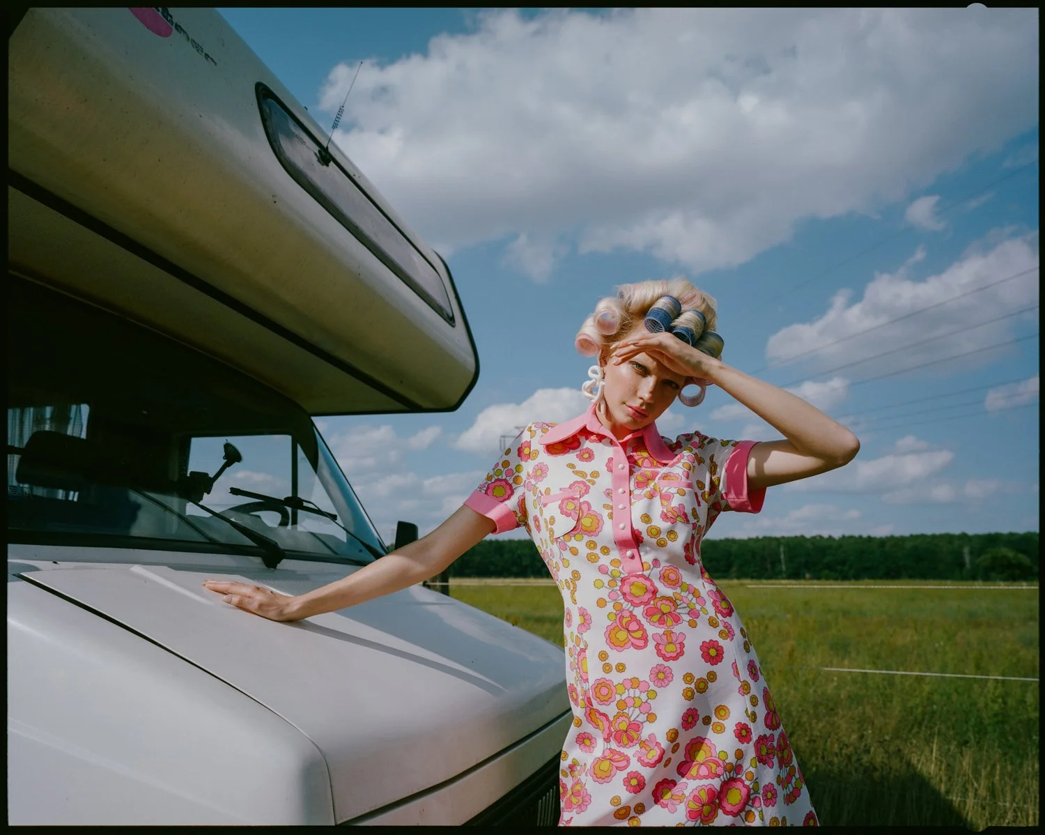 A woman dressed in a vintage floral dress with curlers in her hair, standing next to a white camper van in an open field under a cloudy blue sky, with her hand resting on the van and shading her eyes.