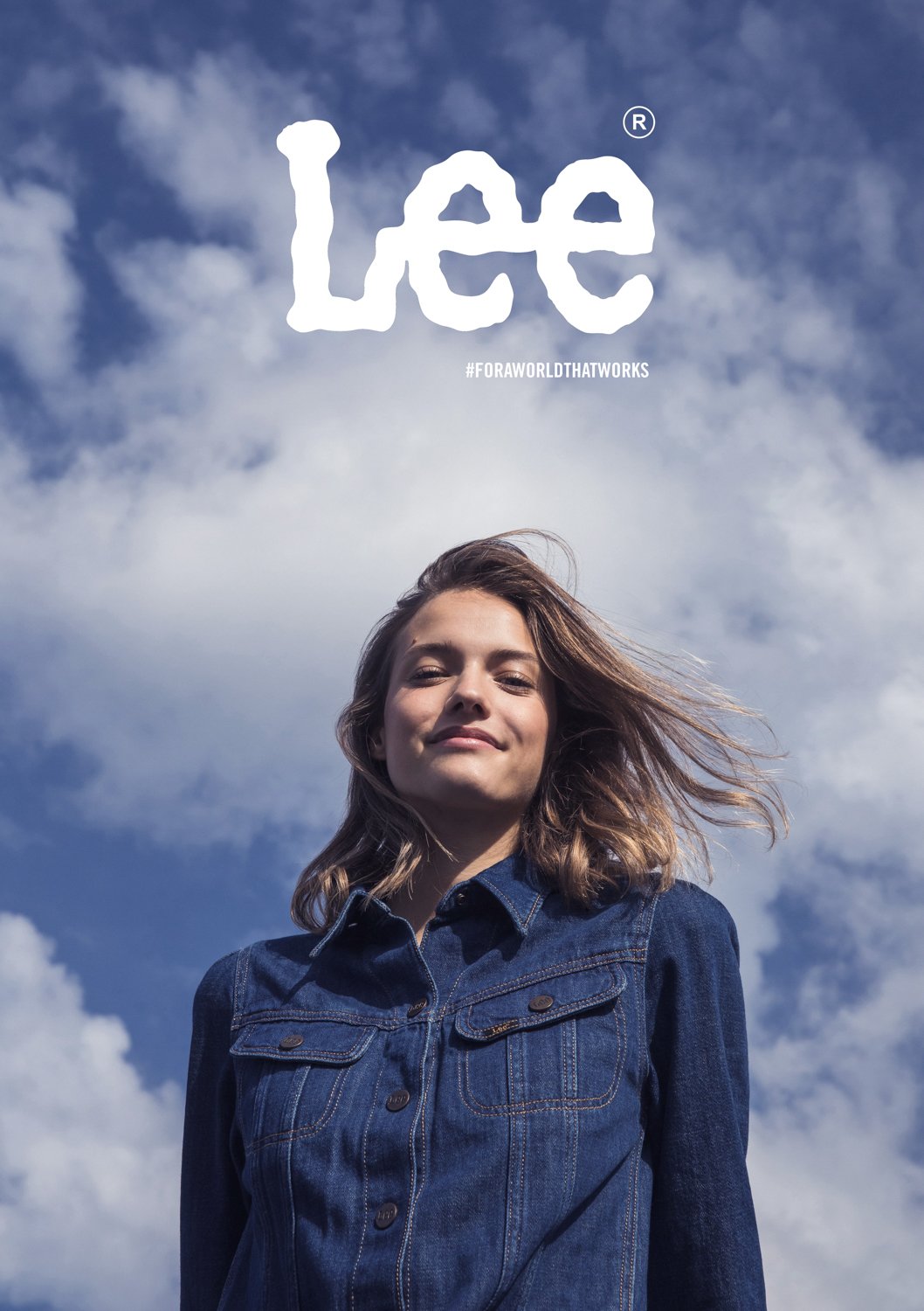 A woman with shoulder-length brown hair wearing a denim jacket standing against a cloudy sky