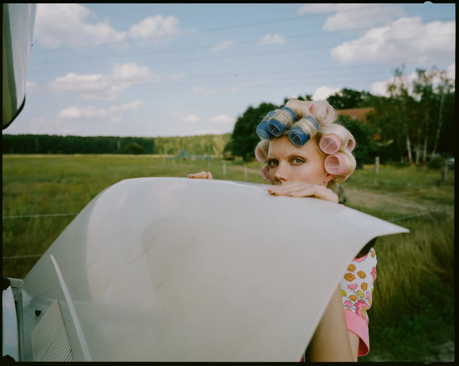 A woman with pink hair rollers peeking over a white object outdoors with a grassy field and a blue sky with clouds in the background.