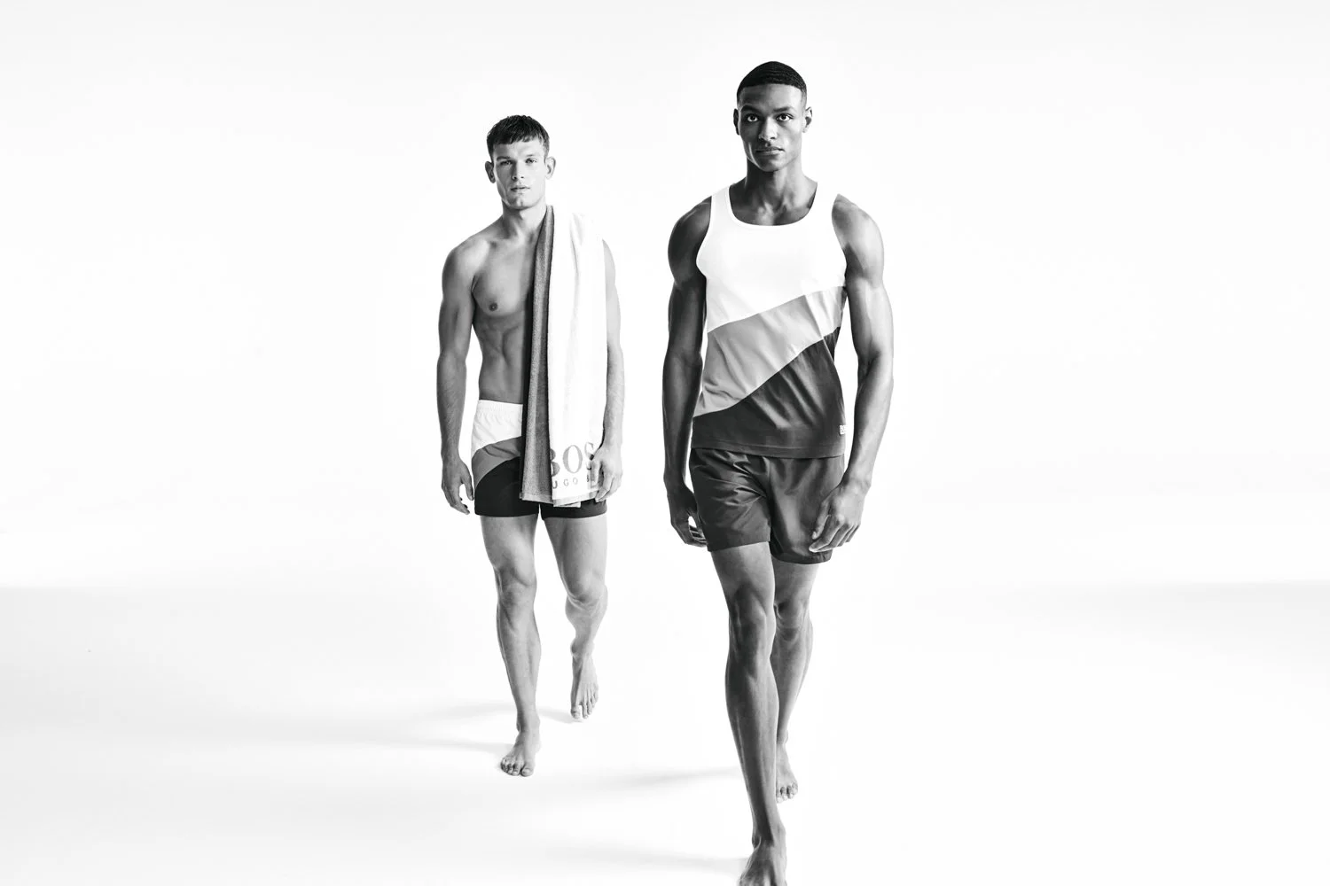 Two male models walking barefoot in athletic wear, one carrying a towel over his shoulder, against a plain white background in black and white.