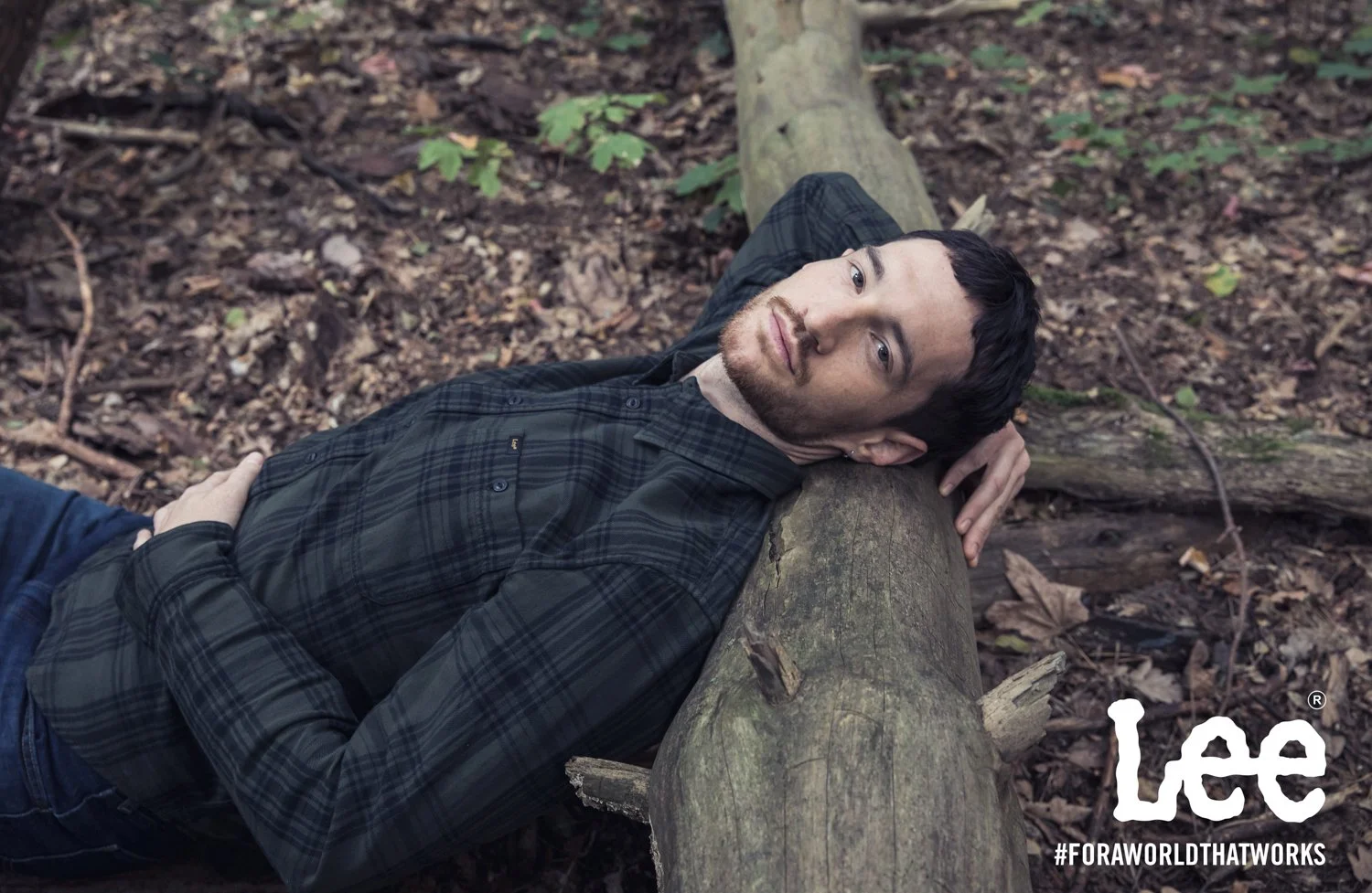 A man with dark hair and light beard lying on a fallen tree in a wooded area, looking up at the camera. He's wearing a dark plaid shirt and blue jeans. The ground is covered in leaves and small branches. The Lee logo and hashtag #FORAWORLDTHATWORKS a