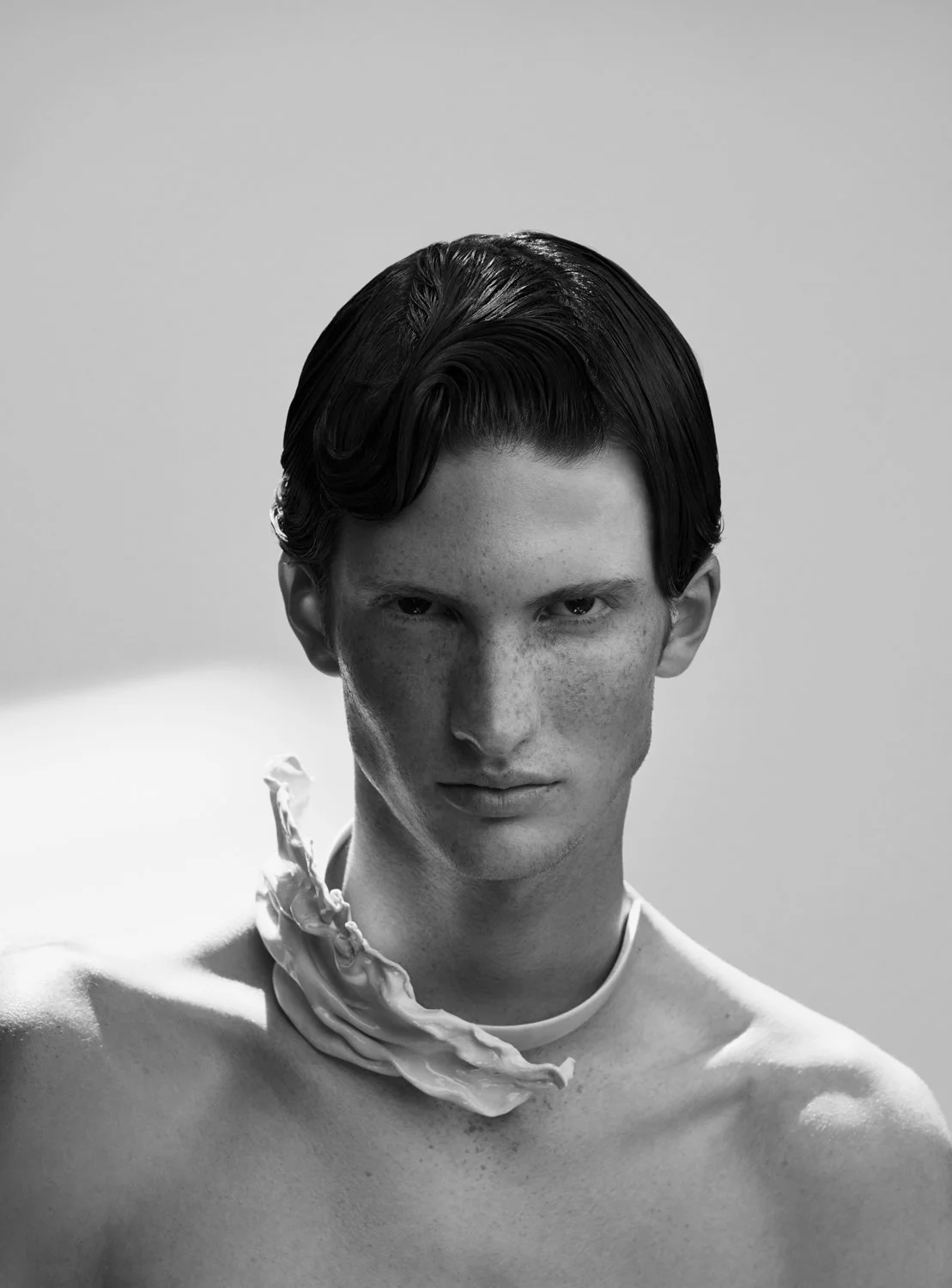 A black-and-white portrait of a young man with dark, wavy hair, serious expression, and a piece of fabric or ribbon wrapped around his neck, against a plain background.
