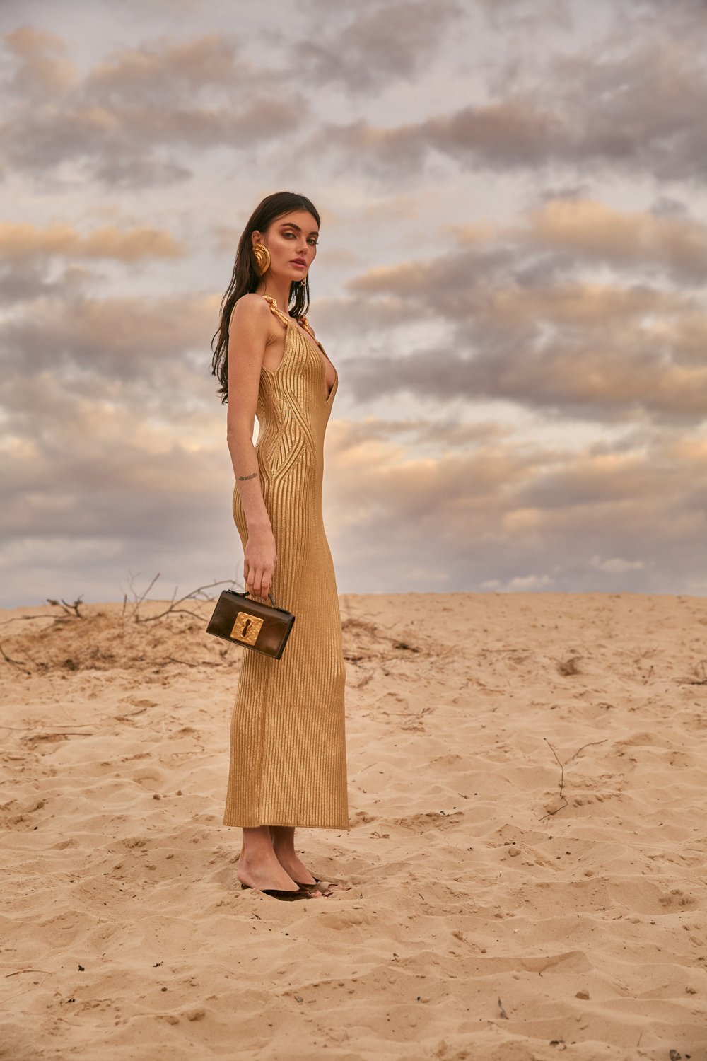 A woman in a gold, sleeveless, long dress stands on a sandy desert landscape under a cloudy sky, holding a black handbag with a gold clasp, wearing high heels and gold jewelry.