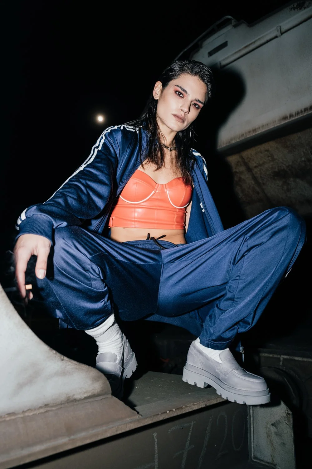 A woman in sporty attire sitting on a ledge at night, wearing a blue track jacket and pants, white shoes, and an orange top, with dark wet hair and a confident expression.