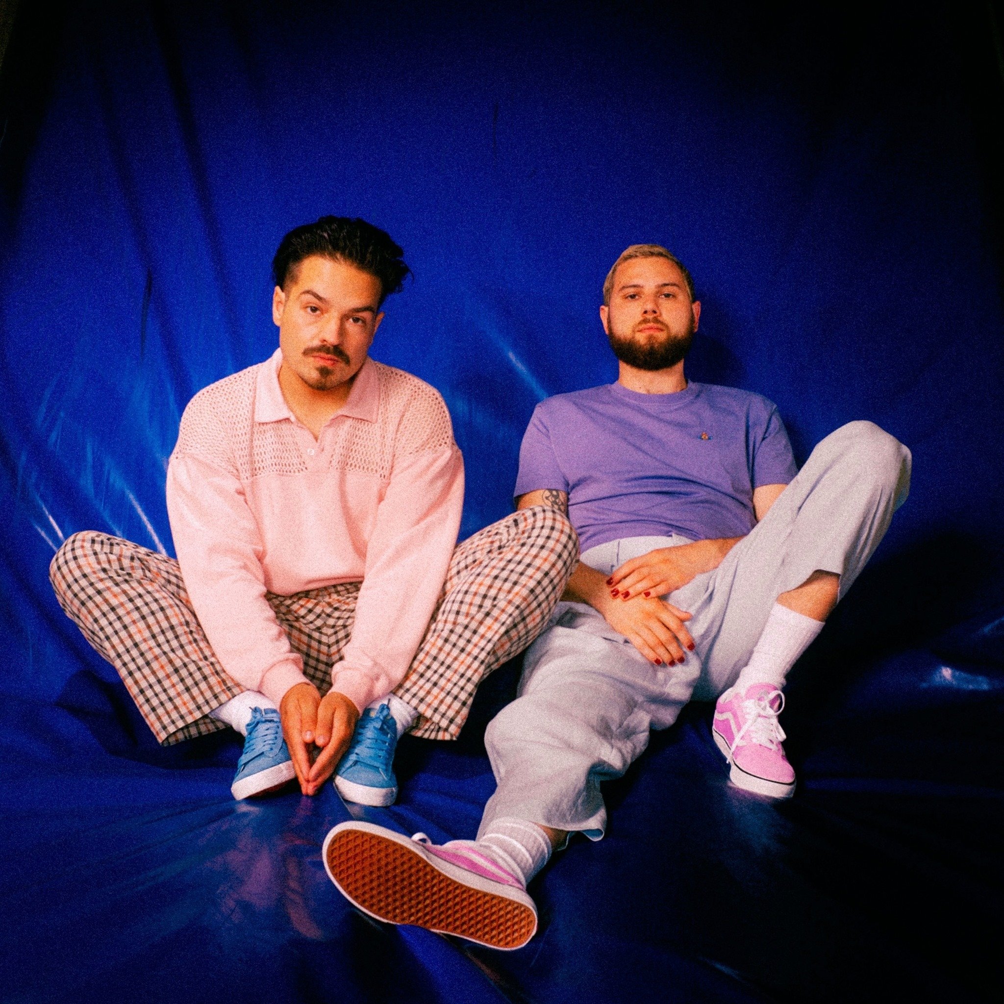 Two young men sit on a blue surface with a blue background, dressed casually, with neutral expressions. One wears a pink sweater and plaid pants, the other wears a purple t-shirt and gray pants.