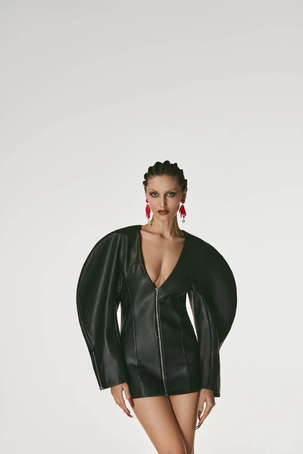 Fashion model wearing a black leather dress with exaggerated shoulder sleeves and red earrings, standing against a plain white background.