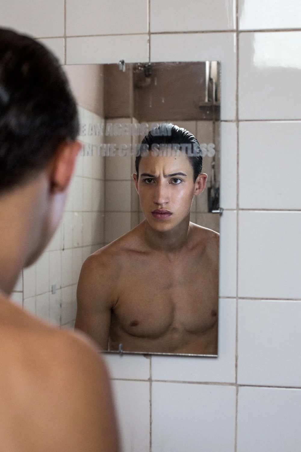 A shirtless man looking at his reflection in a bathroom mirror with a worried or serious expression.