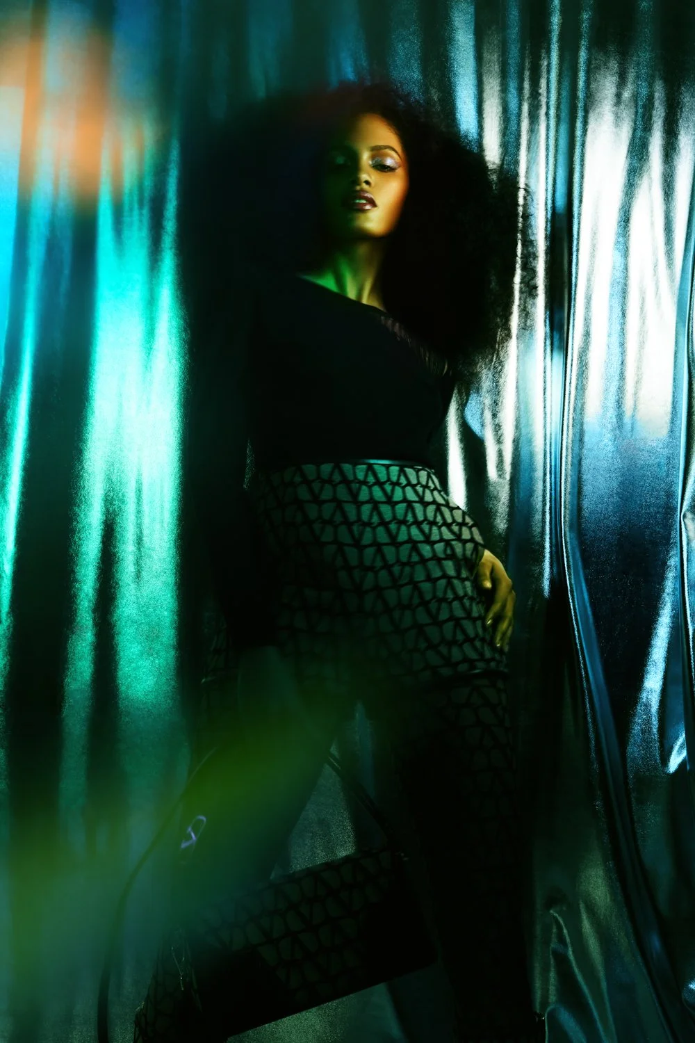 A woman with voluminous curly hair leaning against metallic curtains, illuminated by moody green and blue lighting, wearing a black top and patterned pants.