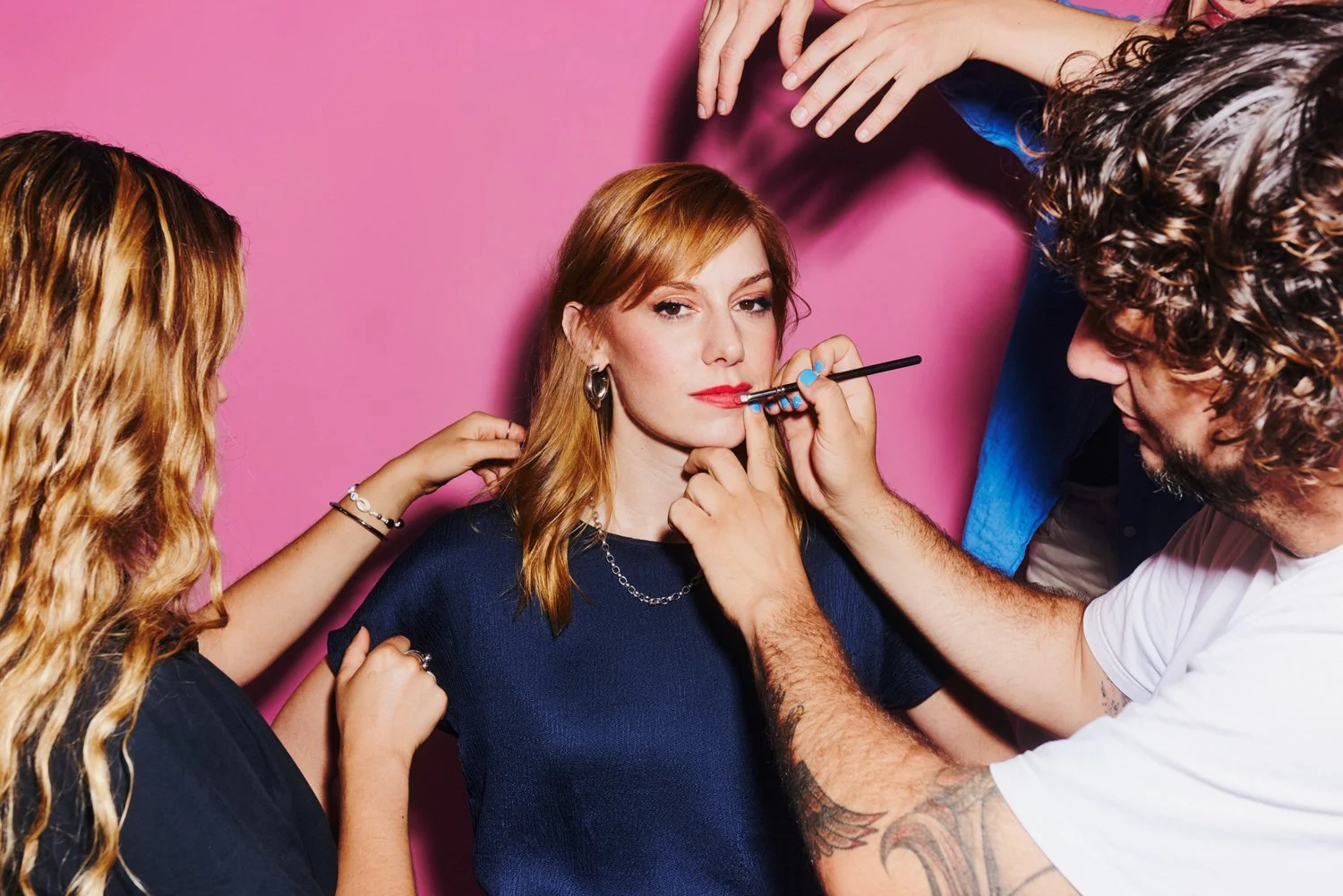 A woman getting her makeup done by a makeup artist while another person assists on her other side, against a pink background.