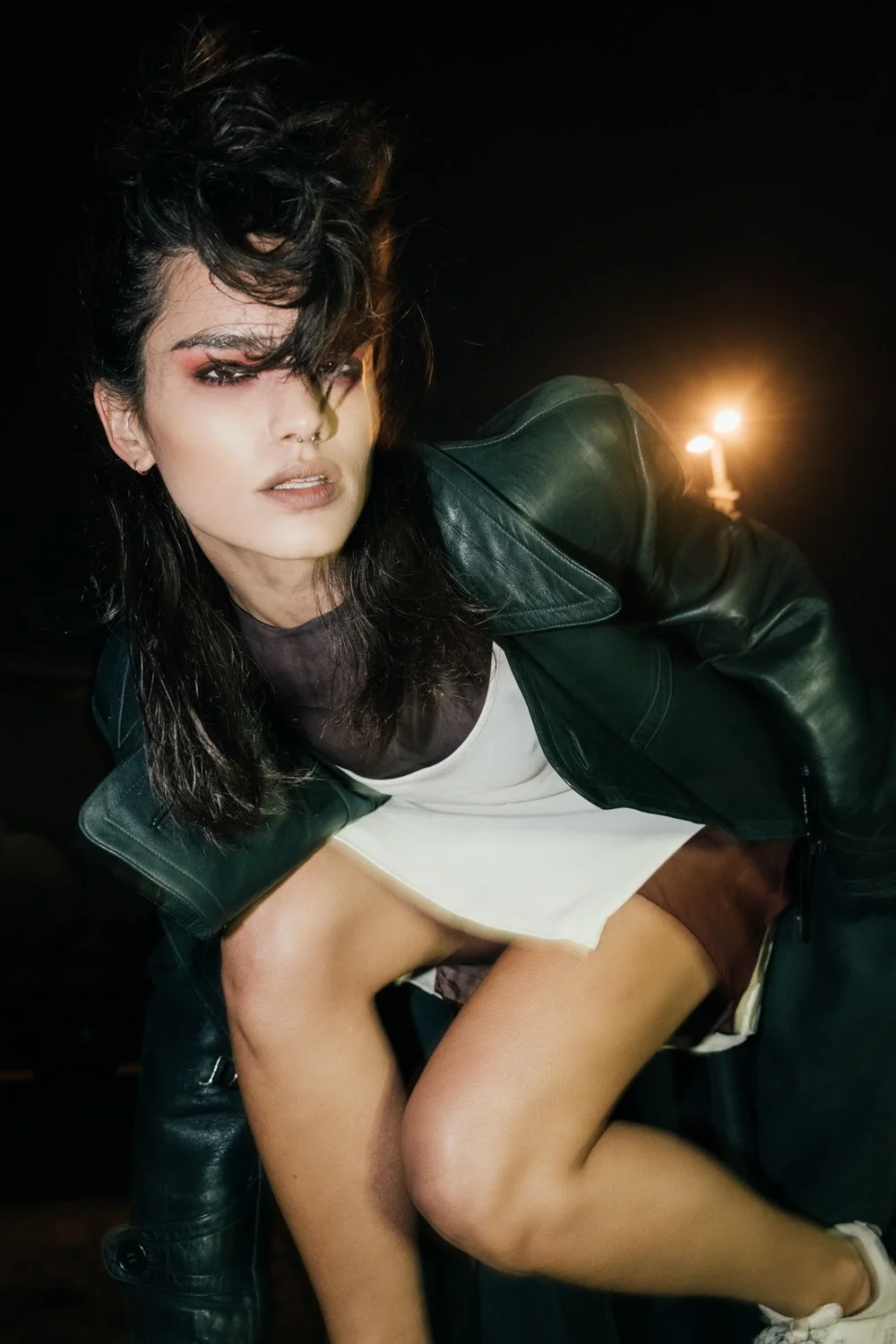 A woman with dark hair, wearing a black leather jacket, white dress, and makeup, outdoors at night with a lit candle in the background.