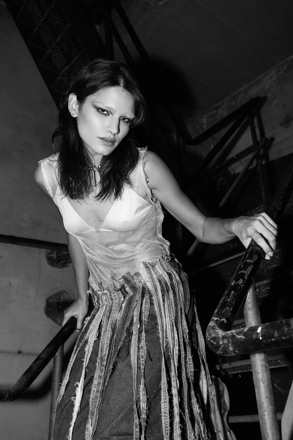 A young woman with dark hair and a piercing, wearing a sleeveless top and striped skirt, leaning on a staircase railing in an industrial setting.