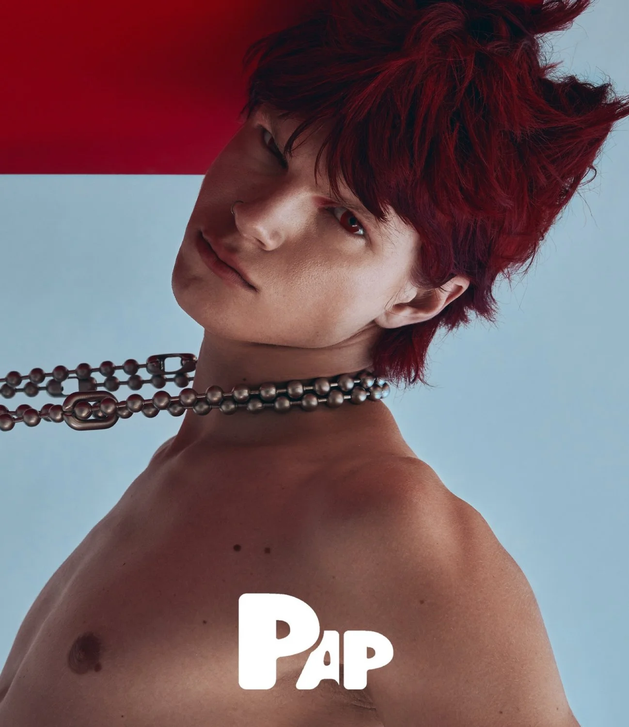 Close-up of a young man with red hair and a septum piercing, looking at the camera, wearing a pearl necklace, with a red and light blue background, and the word 'PAP' at the bottom.