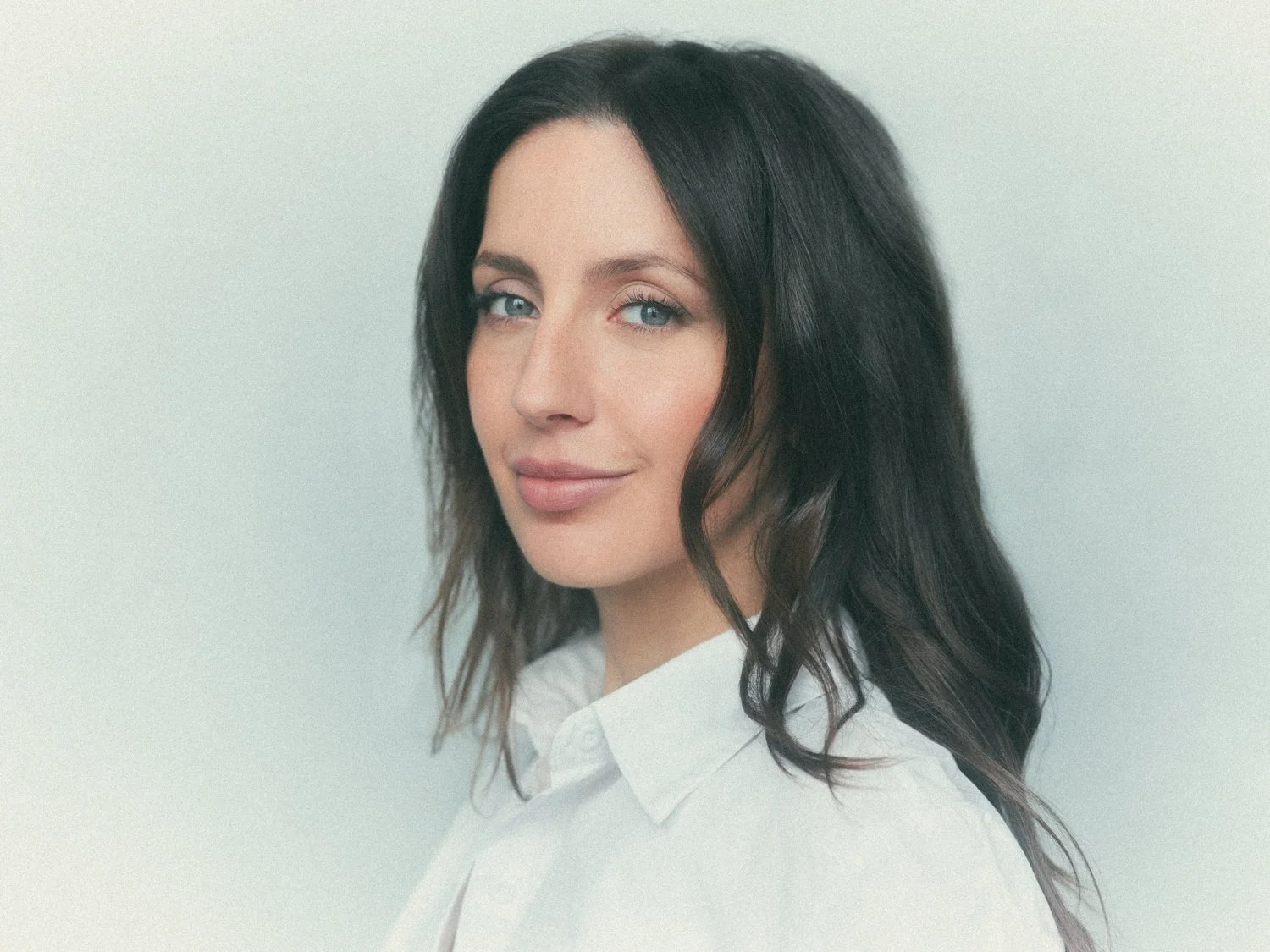 A woman with long dark hair and blue eyes looking at the camera, wearing a white button-up shirt against a light background.