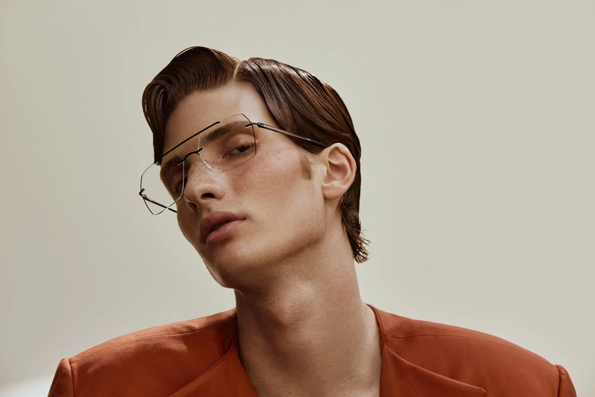 Close-up portrait of a young man with fair skin, red hair, and freckles, wearing stylish glasses and an orange blazer, looking sideways with a neutral expression against a plain background.