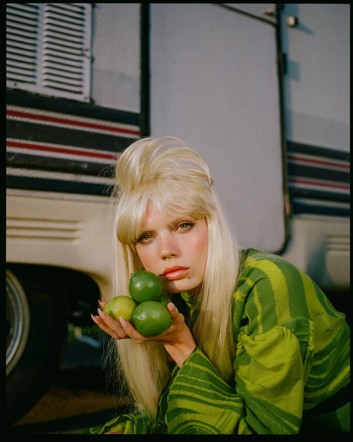 A woman with long blonde hair and green eyes holding four green limes, leaning against a vehicle with a striped pattern.