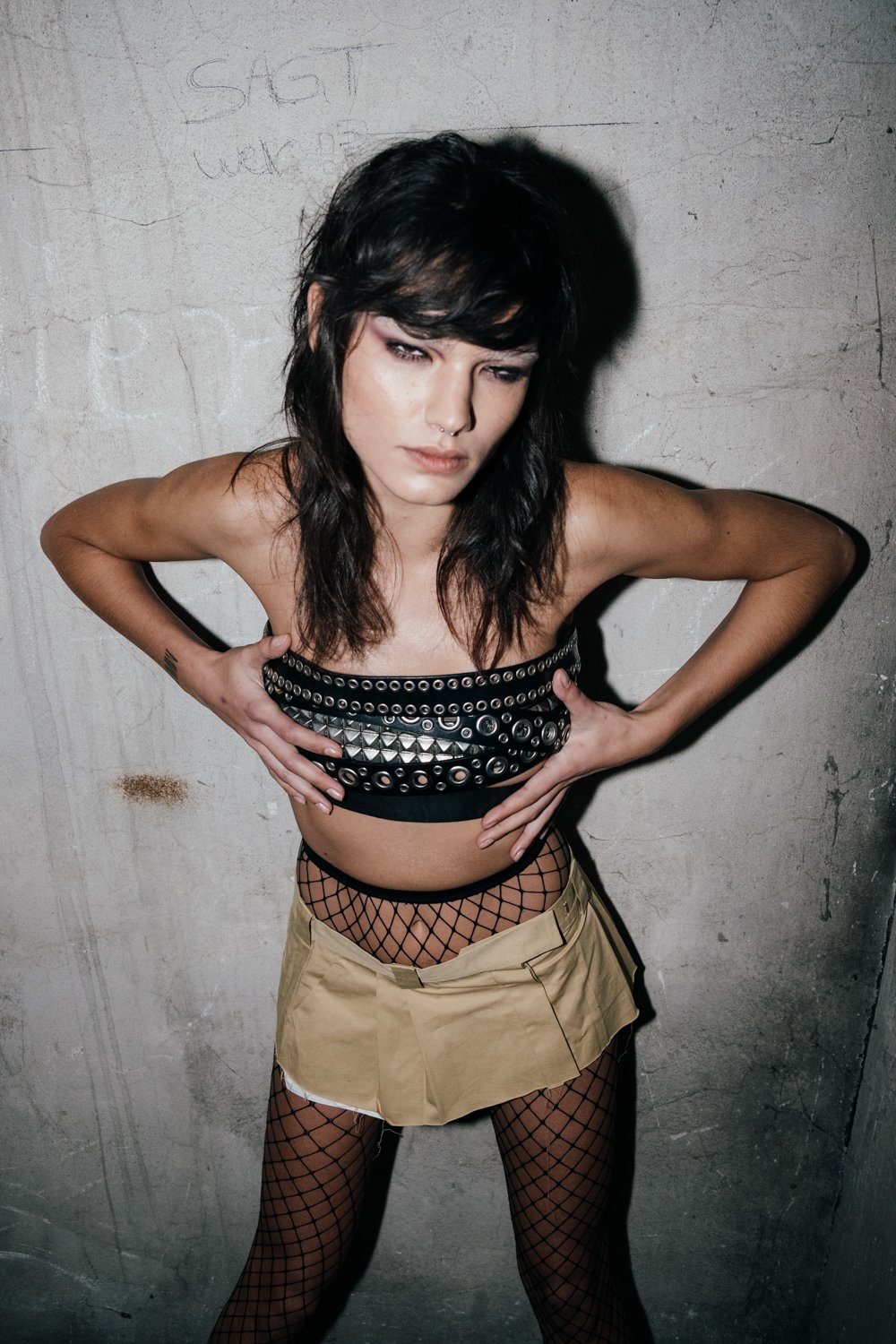 A woman with dark, tousled hair and a serious expression, wearing a black studded top, fishnet stockings, and a tan mini skirt, standing against a gray concrete wall.