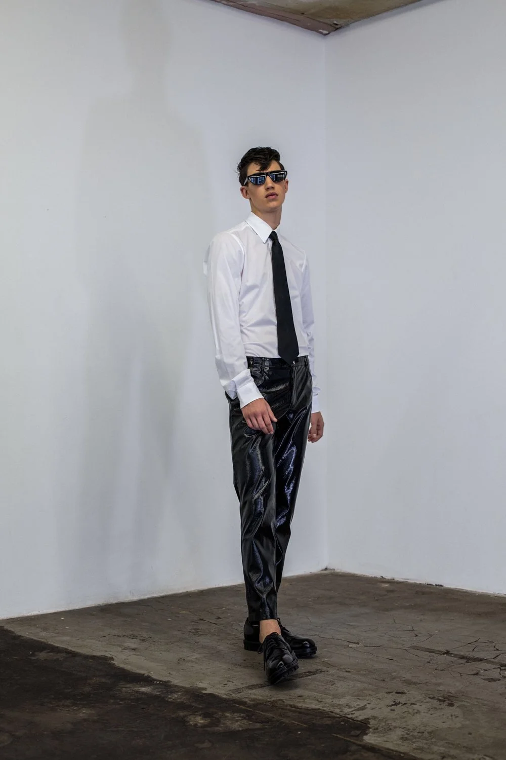 Young man wearing a white dress shirt, black tie, black shiny pants, black shoes, and sunglasses, standing in a corner of a white room with a concrete floor.