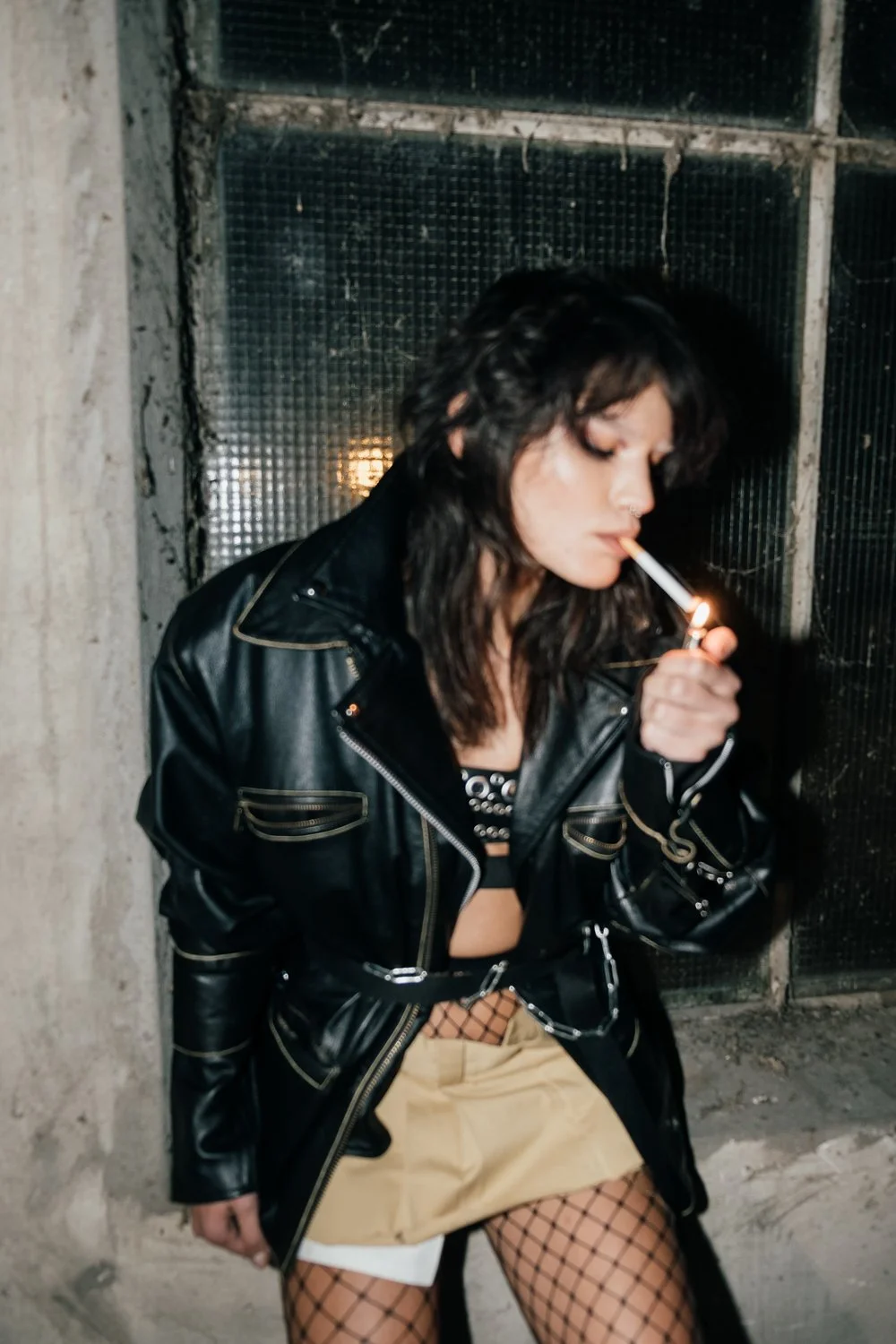 A young woman with dark curly hair wearing a black leather jacket, fishnet stockings, and beige shorts, standing against a gritty urban wall while lighting a cigarette at night.