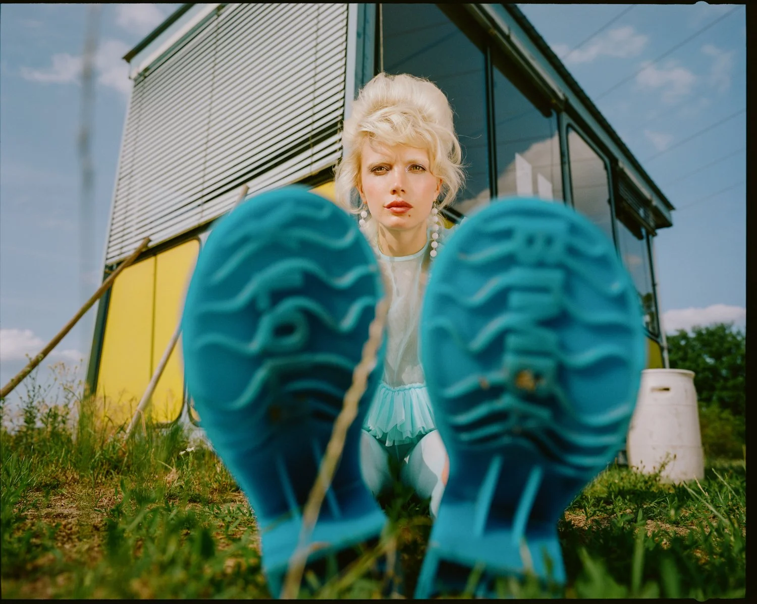 A woman with blonde hair and earrings is looking down at the camera with her feet in blue shoes in the foreground, posed on a grassy area in front of a yellow and black bus, with the sky and some bushes in the background.