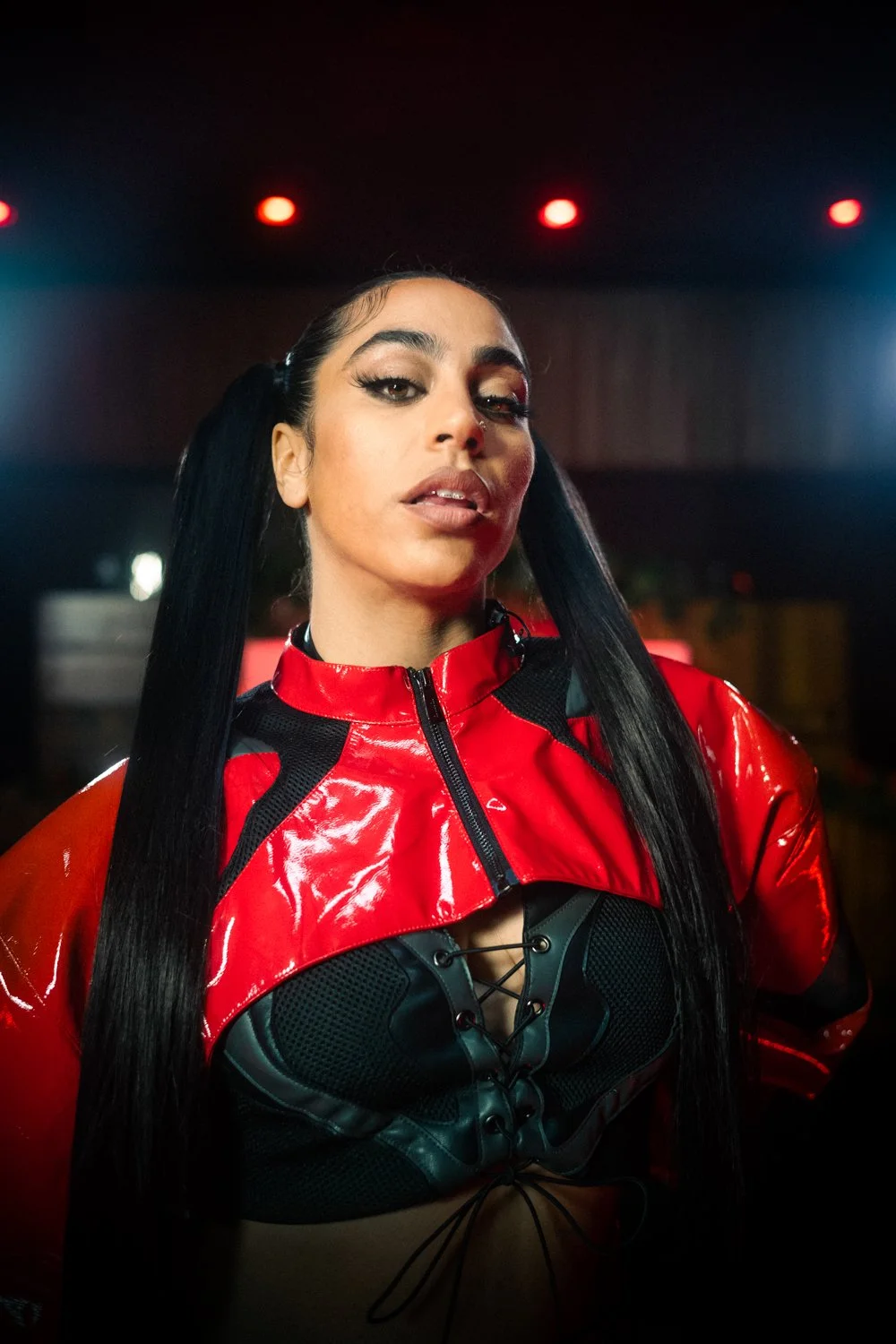 A woman with long black hair, wearing a shiny red and black jacket, posing in a dimly lit indoor setting.