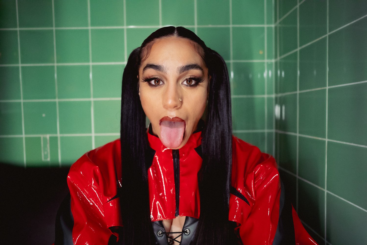 Young woman with long black hair, wearing a shiny red jacket, sticking out her tongue, in front of green tiled wall.