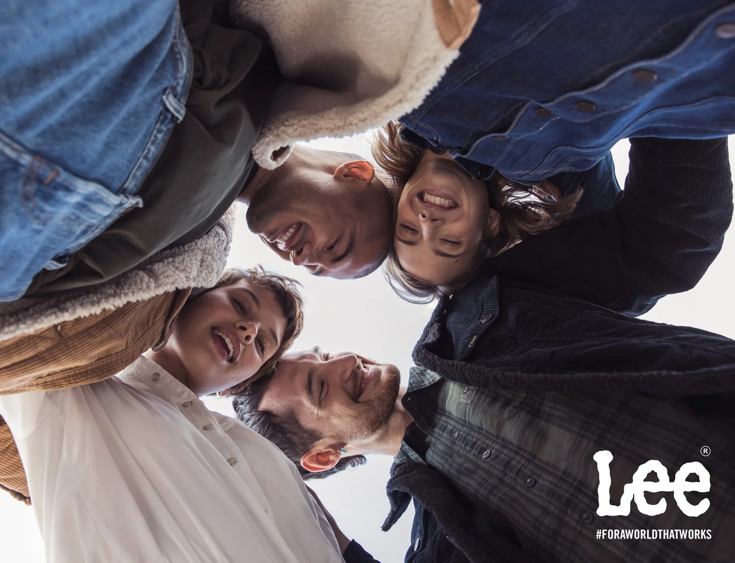 Group of five diverse people standing in a circle with their heads together, smiling and looking down at the camera. It appears to be an outdoor setting.