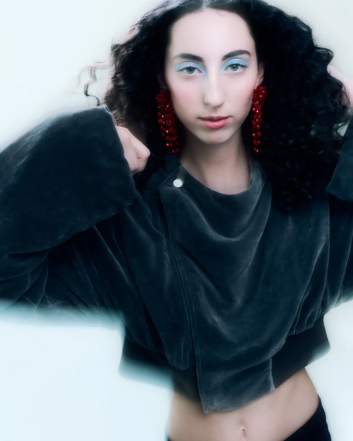 A woman with dark curly hair, blue eyeshadow, and red earrings, wearing a black velvety jacket, looking directly at the camera.