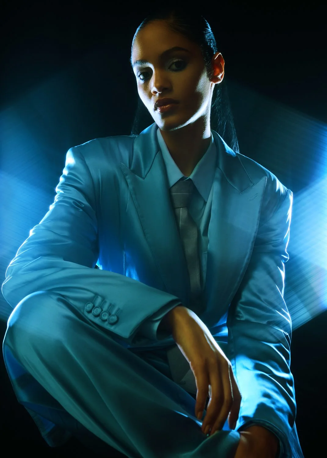 Woman in a light blue suit with sombrero collar and tie, sitting with her leg crossed, in front of a dark background with blue lighting.