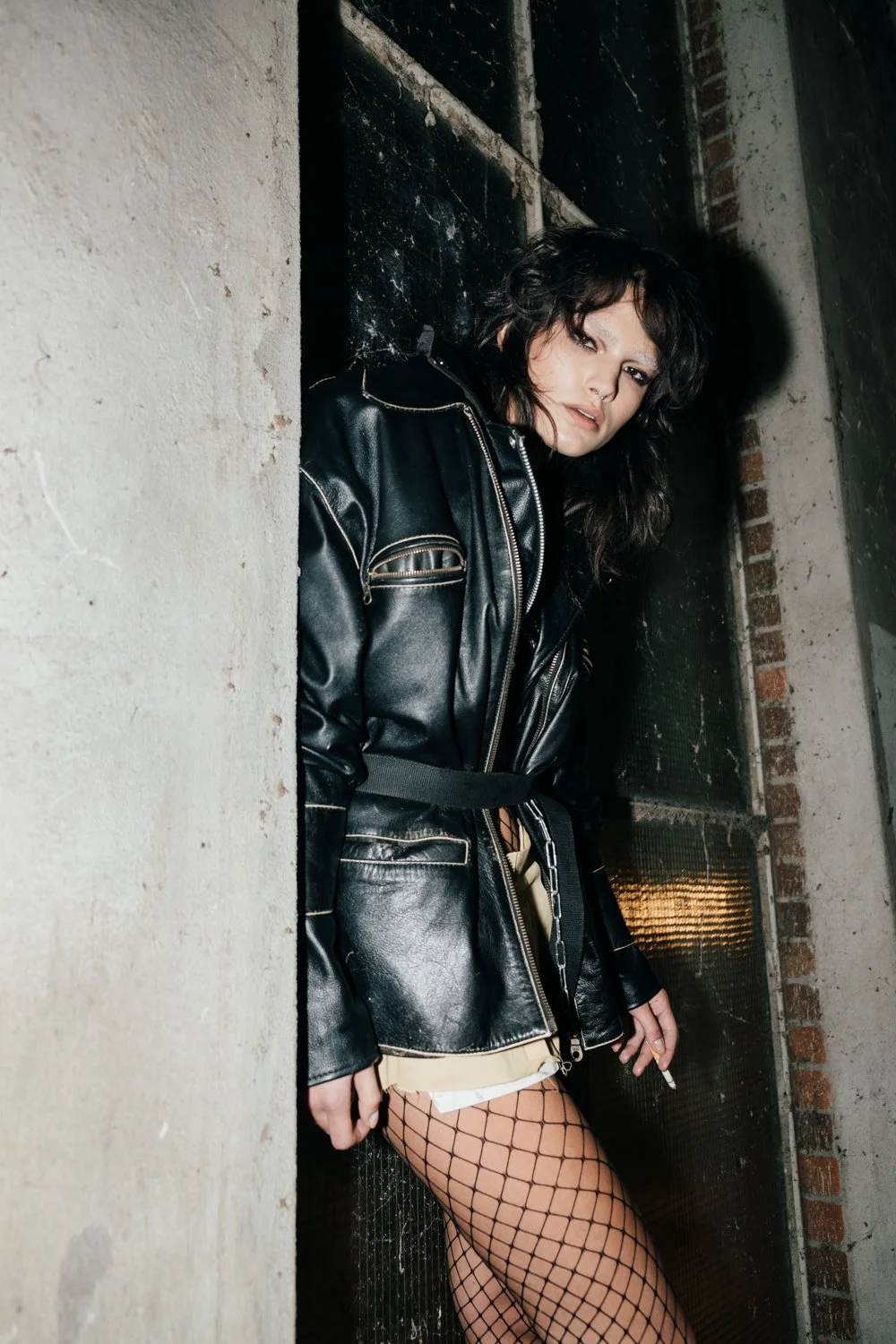 Woman with dark wavy hair in a black leather jacket and fishnet stockings leaning against a wall in an industrial-style setting.