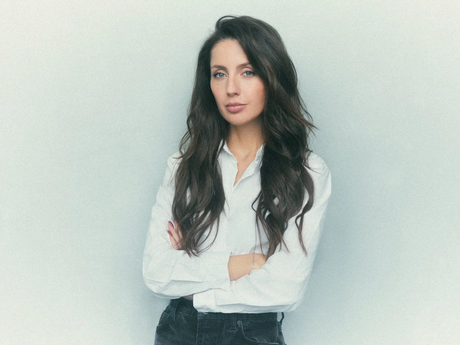 A woman with long, wavy dark hair standing against a plain light-colored wall, crossing her arms and looking directly at the camera.