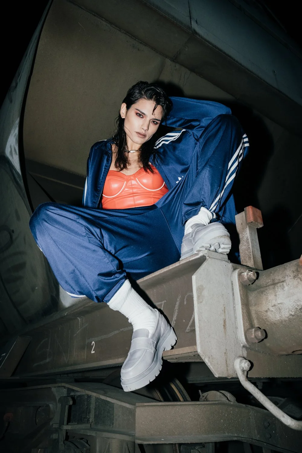 A woman with dark hair wearing a blue Adidas tracksuit, a red top, white sneakers, and white socks, sitting on a metal structure in a parking garage with a serious expression.