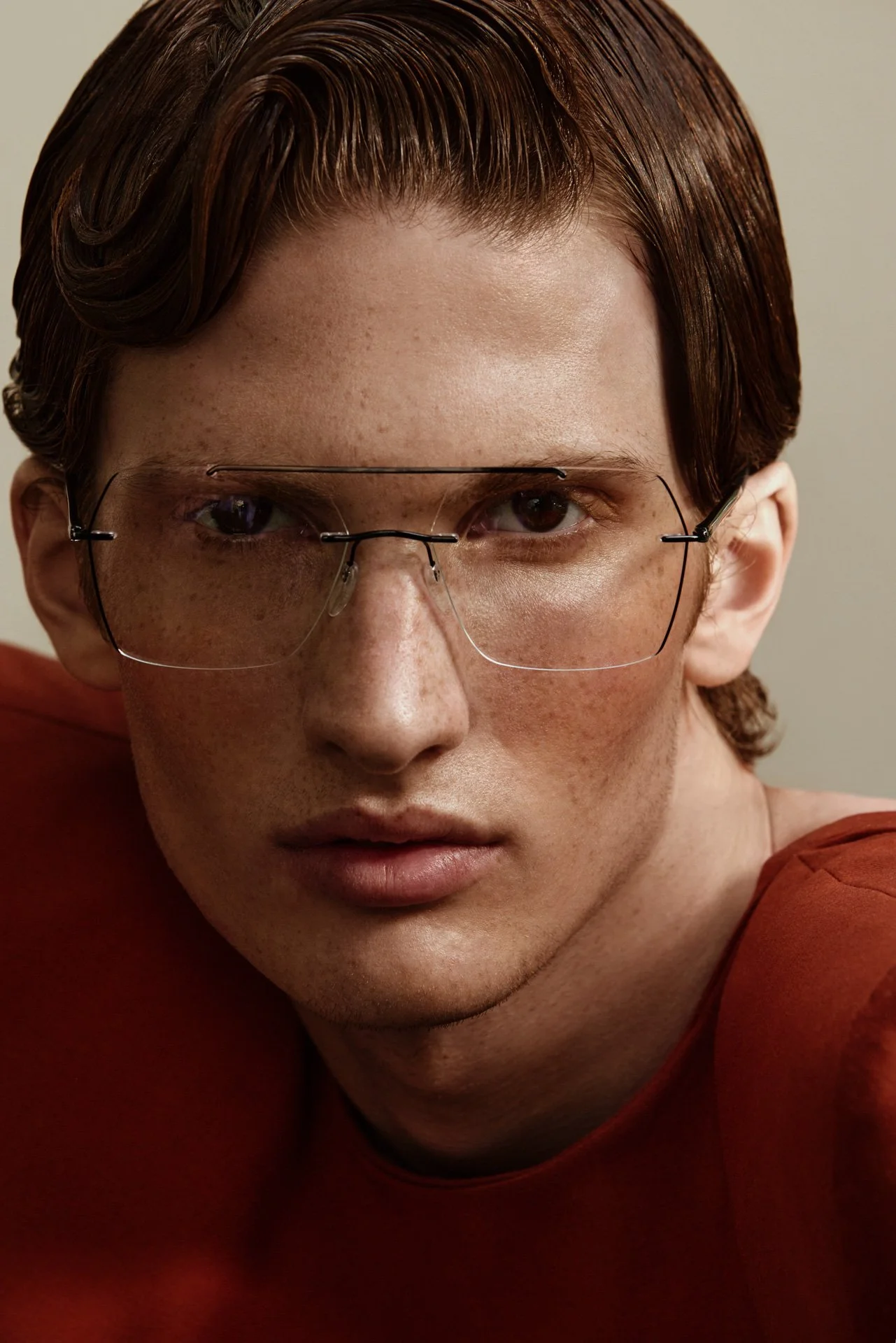 A close-up portrait of a young man with red hair, freckles, and fair skin, wearing large, clear glasses and a red top, looking directly at the camera.