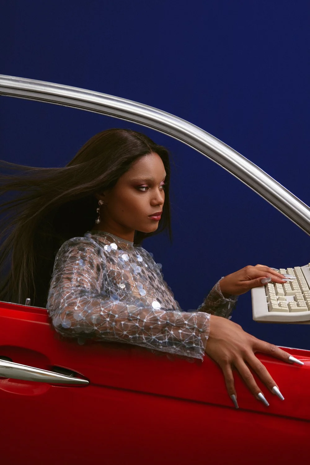 A woman with long dark hair, wearing a translucent top with shiny circular embellishments, sitting in a red car. She is using a keyboard that is held by an unseen person or device, with a dark blue background.