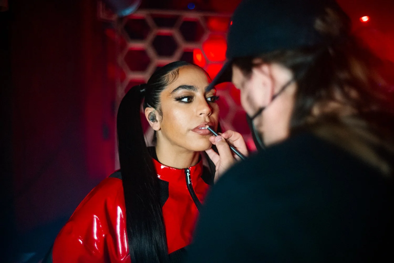 A woman with long dark hair and makeup being assisted by a person in a black cap and glasses in a dimly lit environment with red lighting.