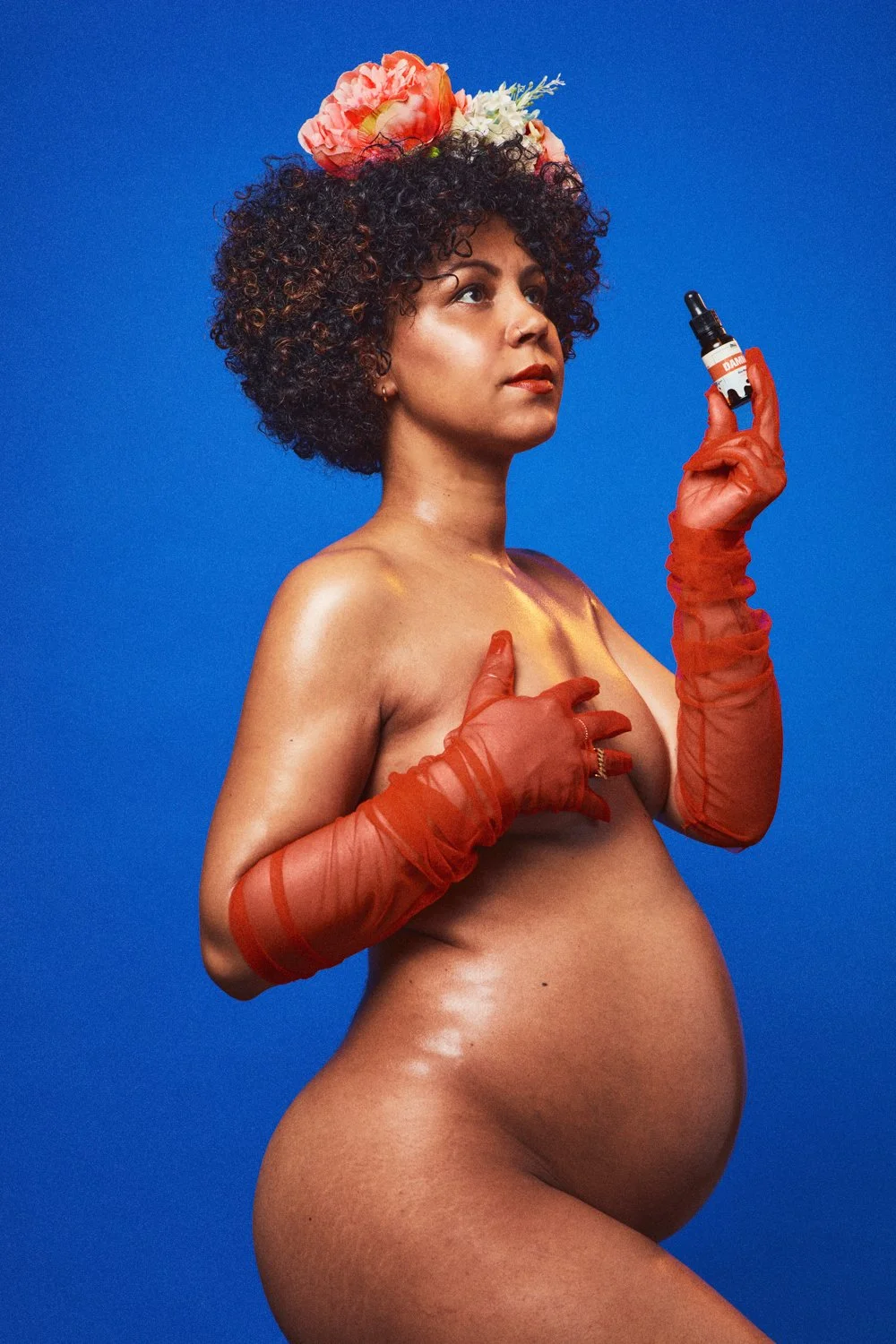 A pregnant woman with dark curly hair, wearing orange sheer gloves and a floral headpiece, holding a skincare dropper bottle, standing against a blue background.