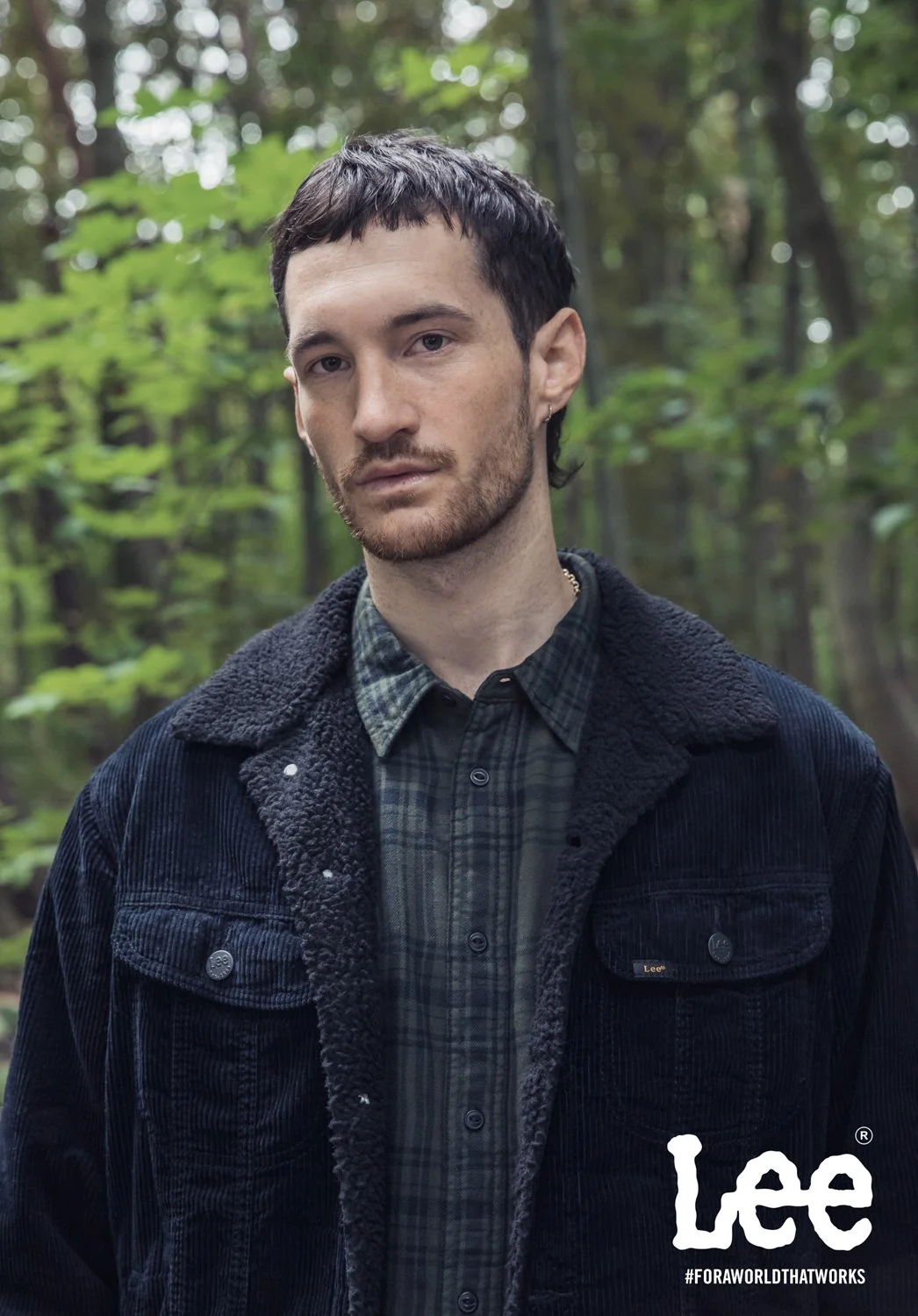 A young man with short dark hair and a beard stands outdoors, wearing a dark cardigan with a fleece collar over a plaid shirt, with a forest in the background.